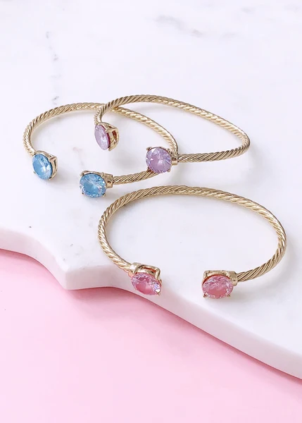 Three gold cuff bracelets with blue, purple, and pink gemstones on white marble