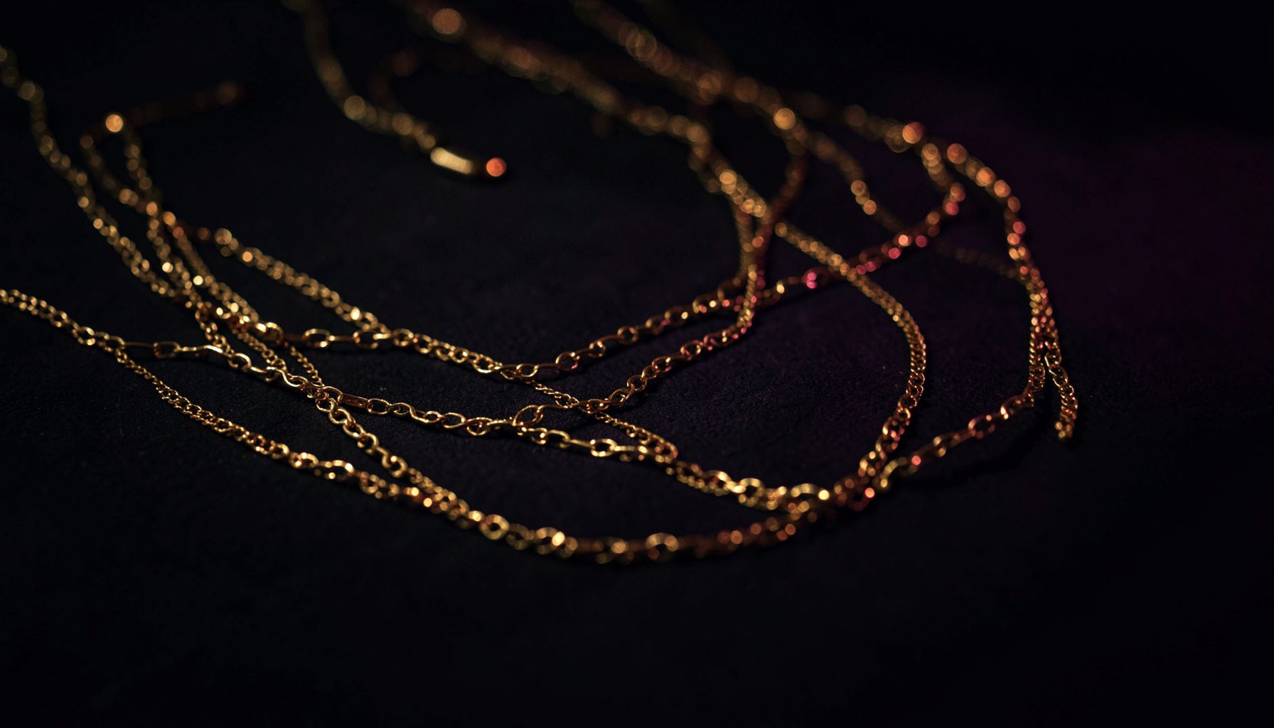 Dark moody photograph of layered gold necklaces resting on a deep charcoal surface, warm amber light catching the metal edges
